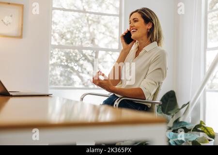 Cheerful businesswoman taking a phone call in her home office. Happy young businesswoman communicating with her business partners while working remote Stock Photo