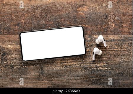 mockup of cell phone placed horizontally with white blank screen and wireless headphones on background of an old wooden table. Game, video streaming s Stock Photo
