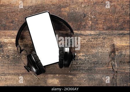 Cellphone mockup display with black wireless headphones on wooden desk background. Copy space. Audio apps, music podcasts books. phone blank screen an Stock Photo