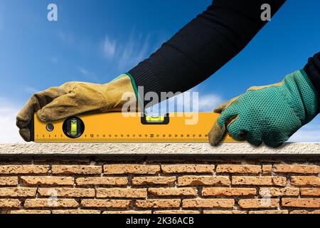 Manual worker with protective work gloves, holding an orange construction spirit level, above a brick and marble wall with blue sky on background. Stock Photo