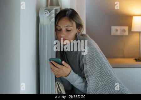 Upset woman leaning on heating radiator searches vacancy for applying for job scrolling smartphone Stock Photo