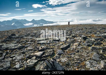 Female hiker coming to the top of stony mountain with clouds and vast arctic landscape in the background. Top of Naite,Sarek National Park, Sweden Stock Photo