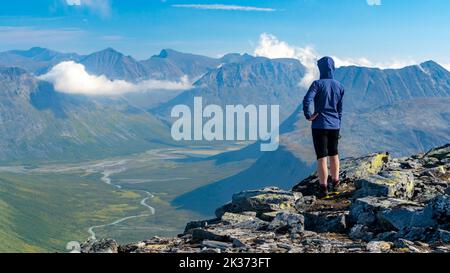 Female hiker in blue jacket overlooking vast arctic mountain landscape from the top of the mountain. Top of Naite, Sarek National Park, Sweden. Hiking Stock Photo
