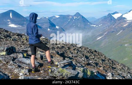 Female hiker in blue jacket overlooking vast arctic mountain landscape from the top of the mountain. Top of Naite, Sarek National Park, Sweden. Hiking Stock Photo