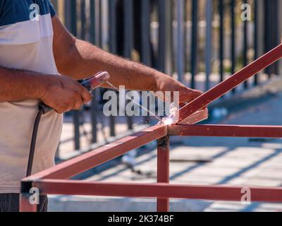 Welder welding two iron profiles at the construction site on a sunny day Stock Photo