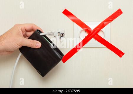 A mans hand holds a wallet with a white cord hanging from it with a plug near an electrical outlet Stock Photo