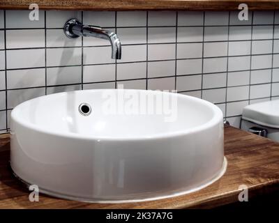 Clean modern loft style bathroom interior with wall-mount stainless steel faucet and white round shape sink basin on brown wooden plank shelf on white Stock Photo