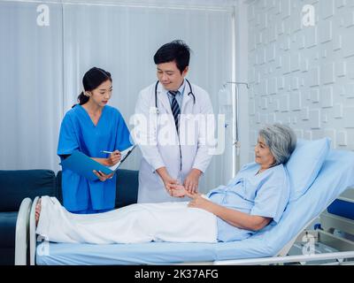 Asian male doctor, professional in white lab coat holding elderly patient's hand for check to examine heartbeat pulse and nursing assistant taking not Stock Photo