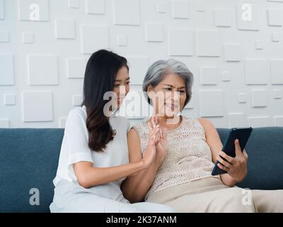 Happy lovely Asian senior woman, mother white hair and beautiful young female, daughter waving greeting at digital tablet screen while sit on couch in Stock Photo