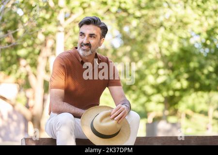 Relaxed middle-aged man holding a hat and looking away while sitting on a bench in a park. Stock Photo