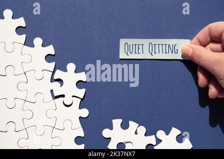 Quiet quitting text, caption. Jigsaw puzzle pieces half assembled and separate. Hand with sticky paper, office sticker.Top view, flat lay, blue white Stock Photo