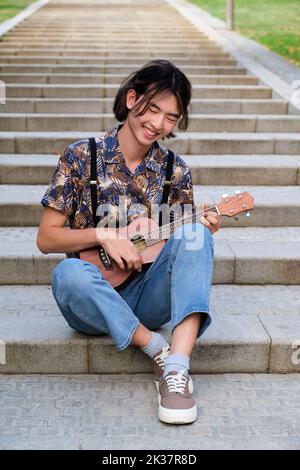 Taiwanese guy smiling and playing acoustic Ukulele guitar sitting on stairs. Stock Photo