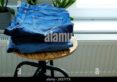 stack of blue denim jeans on stool in bedroom aganst window Stock Photo