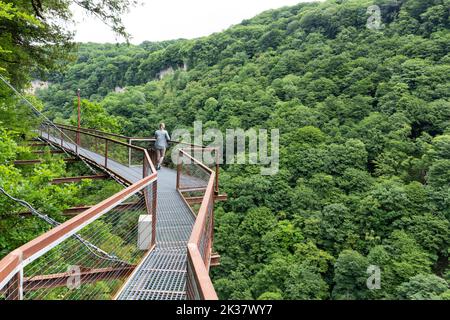Woman traveler in Okatse Canyon in Georgia, standing on hanging metal pedestrian pathway trail above deep precipice, lush vegetation and forests. Stock Photo