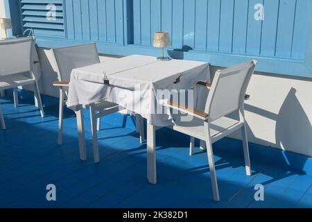 White table and chairs with blue wooden floor and wall as backdrop. Outdoors in sunshine with shadows. Restaurant, no people. Stock Photo