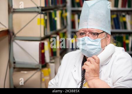 An elderly tired woman doctor working in the reception of a clinic is answering phone calls and making appointments during a big epidemic. Tired eyes Stock Photo