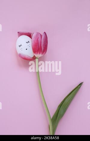 Egg with face sleeping on tulip over pastel pink background. Symbol of picture Easter, tulip with chicken egg. Painted egg face. Easter decorations. Stock Photo