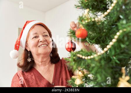Happy senior hispanic woman smiling while decorating a Christmas tree wearing a red Santa Claus beanie. The joy of the holidays. Stock Photo