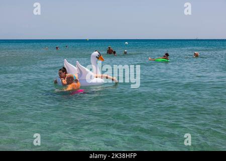 Beautiful view of people swimming in sea. Couple on inflatable white swan on front. Rhodes. Greece. Stock Photo
