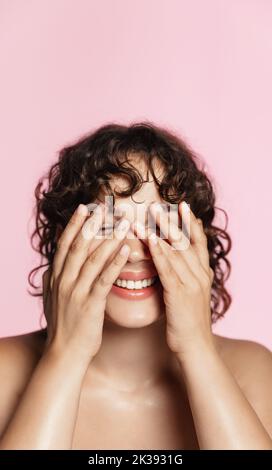 Vertical shot of curly girl washes her face, using cleansing gel treatment from blemishes, better skin tone and no acne, pink background. Stock Photo