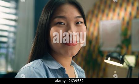 Attractive asian woman looking and smiling at camera while sitting at home office at the table near computer and having remote work Stock Photo