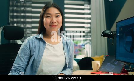 Female asian 3D designer sitting at the table near the modern computer and digital tablet, smiling and looking at camera while working on 3D project remotely from home. Stock Photo