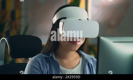 Woman in VR headset sitting at the table at home and working remotely on computer or surfing the internet in spare time. Virtual reality. Stock Photo