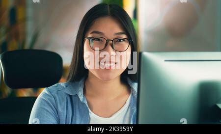 Asian woman in glasses looking at screen of computer while working on 3D modeling project or design remotely from home or surfing the internet in spare time. Freelance Stock Photo