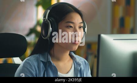 Asian woman in headphones sitting at the table at home and browsing the internet on computer on weekend or working on project remotely Stock Photo