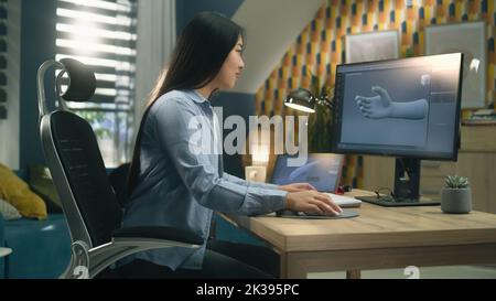 Asian woman sitting on chair at the table at home office and making 3D prototype of human hand in professional program for 3D modelling, using modern computer and digital tablet. Stock Photo