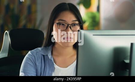 Asian woman in glasses looking at screen of computer while working on 3D modeling project or design remotely from home or surfing the internet in spare time. Freelance Stock Photo