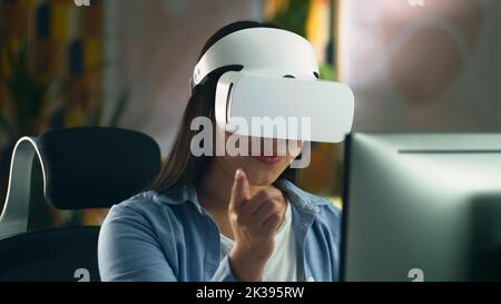 Woman in VR headset sitting at the table at home and working remotely on computer or surfing the internet in spare time. Virtual reality. Stock Photo