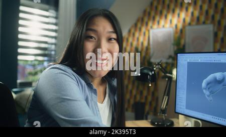 Surprised asian woman rejoicing and looking at camera while sitting at the table at home office and working remotely on 3D modeling project on computer Stock Photo