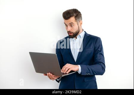 Shocked surprised caucasian bearded business man, in a suit, programmer, IT specialist, holding opening laptop in hands, looks in amazement at the screen, standing on isolated white background Stock Photo