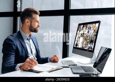 Online virtual meeting. Successful caucasian businessman, sitting in office, having video conversation with group of multiracial partners, discussing company future and perspectives, takes notes Stock Photo