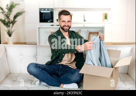 Handsome positive caucasian man opening big carton box while sits on sofa, unpacking internet store order at home, feel satisfied with fast delivery service and the goods which received, smile happily Stock Photo