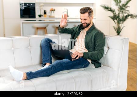 Positive handsome caucasian man, in stylish casual wear, having online video conference with friends or colleagues by laptop at home while sitting on comfortable sofa, waves hand, smiling friendly Stock Photo
