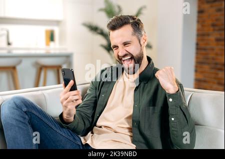 Joyful lucky attractive caucasian man in casual wear, sitting on a couch in cozy living room, using his smart phone, reading good message or news, gesturing with fist, smiling. Digital technology Stock Photo