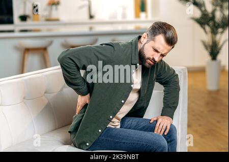 Unhappy caucasian man, dressed in casual wear, sitting on sofa in living room, experiencing acute pain in the back, lower back, has chronic joint problems. Problems with the back, spine, sciatica Stock Photo