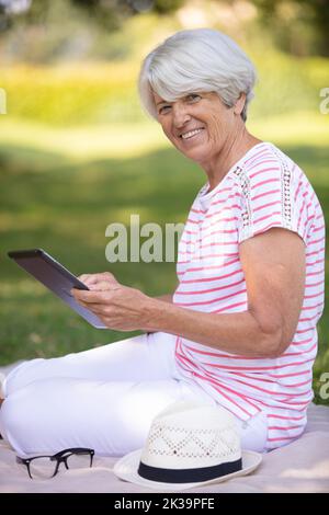 senior woman sits alone on grass using a digital tablet Stock Photo