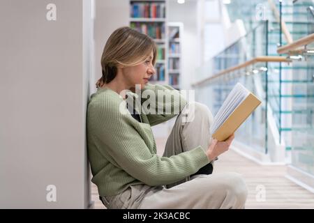 Middle aged student sitting on floor reading book with interesting plot in library. Education. Stock Photo