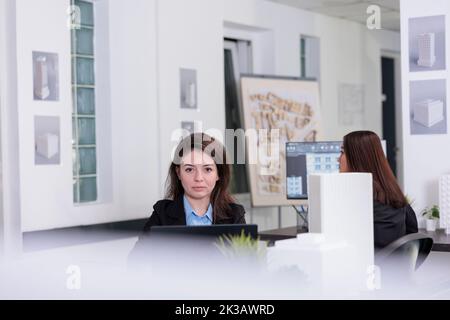 Woman working on laptop in coworking space, looking at camera. Busy employee in architecture office at workplace desk portrait, real estate worker developing small project plan. Stock Photo