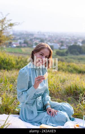 A woman in a long summer dress with short hair sitting on a white blanket with fruits and pastries and eating cheese. Concept of having picnic in a ci Stock Photo