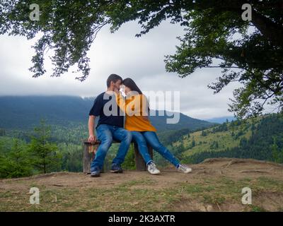 Young man and woman admiring breathtaking view while sitting on bench in the mountains. Lovely couple in casual clothes sit under large tree with fore Stock Photo