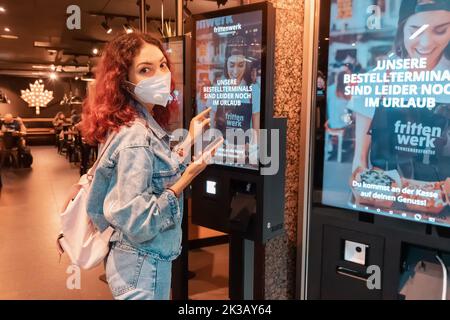 21 July 2022, Dusseldorf, Germany: Woman customer wearing face mask uses a terminal or self-service kiosk to order at a fast food restaurant. Stock Photo
