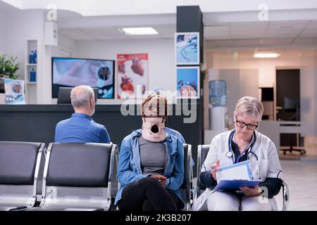 Portrait of patient with neck collar foam sitting in waiting room at clinic doing consultation with senior doctor. Woman wearing cervical brace after accident injury talking to medic about healthcare. Stock Photo