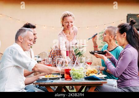 One adult blonde woman smiling and serving plate with salad on table, diverse friends sitting around and having fun. Lifestyle concept. Stock Photo