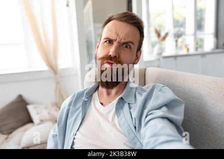 Bearded happy young man smiling and taking selfie photo while sitting on sofa at home. Stock Photo