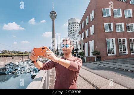 Happy man travel blogger takes selfie pictures on smartphone of the famous Dusseldorf TV tower from the Media Harbor canal in the post industrial dist Stock Photo