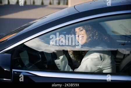 Beautiful businesswoman with closed eyes sitting in electric car and smiling while using vehicle autopilot system. Pretty woman holding steering wheel and sleeping while sitting on driver seat in car. Stock Photo